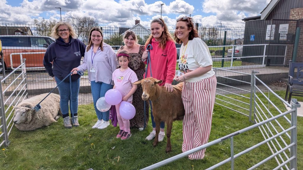 Families and volunteers gathered around a friendly goat and sheep in a mobile farm pen at an accessible farm morning, a child holding balloons